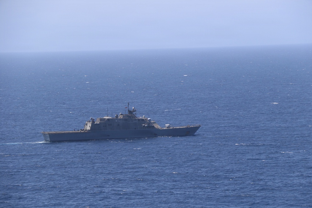 A gray naval warship, consistent with a US Navy Littoral Combat Ship (Freedom-class or Independence-class), is underway on open blue ocean waters. The vessel is photographed from an elevated or distant vantage point, showing its full profile against a hazy blue sky. The ship appears to be on a routine patrol or transit mission.