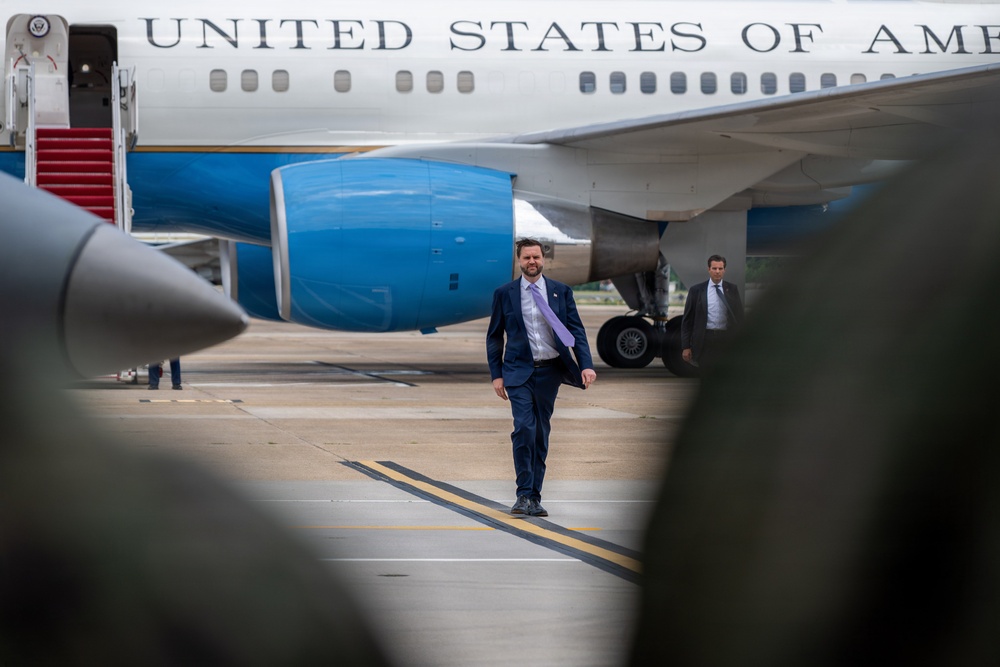 A man in a navy blue suit with a light purple tie walks across an airport tarmac in front of a large aircraft bearing the text 'UNITED STATES OF AME' (partially visible), consistent with a U.S. government aircraft. A second man in a dark suit is visible in the background near the aircraft. The scene appears to be an official arrival or departure at a military or government airfield.