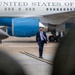 A man in a navy blue suit with a light purple tie walks across an airport tarmac in front of a large aircraft bearing the text 'UNITED STATES OF AME' (partially visible), consistent with a U.S. government aircraft. A second man in a dark suit is visible in the background near the aircraft. The scene appears to be an official arrival or departure at a military or government airfield.