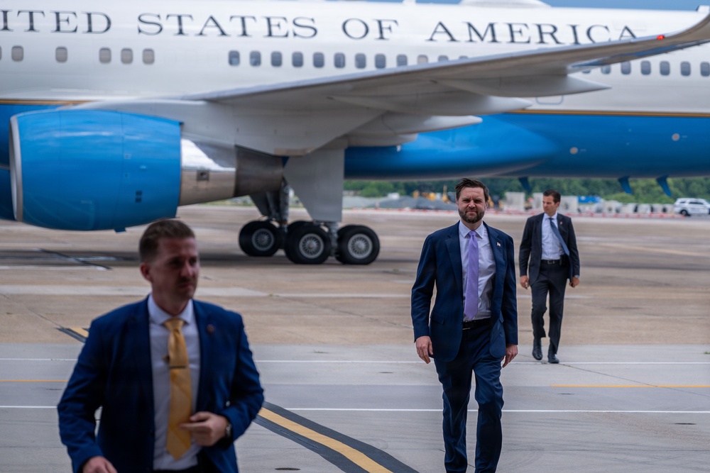Three men in business suits are walking on an airport tarmac in front of a large aircraft bearing the text 'United States of America' on its fuselage. The aircraft appears to be an official U.S. government plane based on its distinctive white and blue livery. The men appear to be departing or arriving at the aircraft.