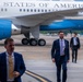 Three men in business suits are walking on an airport tarmac in front of a large aircraft bearing the text 'United States of America' on its fuselage. The aircraft appears to be an official U.S. government plane based on its distinctive white and blue livery. The men appear to be departing or arriving at the aircraft.