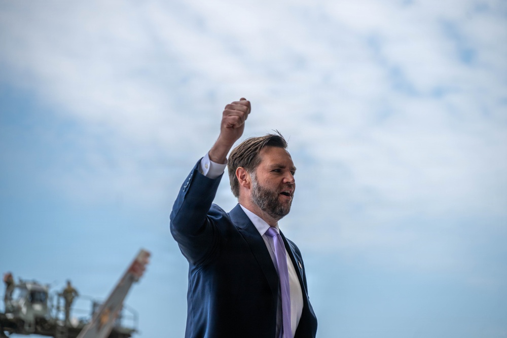 A man in a navy blue suit with a lavender/purple tie is photographed from a low angle against a partly cloudy sky. He has his right fist raised in the air in a triumphant or rallying gesture while appearing to speak. Industrial or mechanical equipment is partially visible in the lower left background.