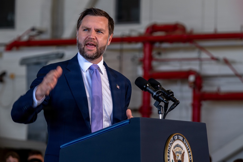 A man in a navy blue suit with a light purple tie is speaking at an official podium bearing a partial seal reading 'PRESIDENT OF THE UNITED STATES.' He is gesturing with his right hand and appears to be delivering a speech in what looks like an industrial or hangar-type facility. An American flag pin is visible on his lapel.
