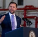 A man in a navy blue suit with a light purple tie is speaking at an official podium bearing a partial seal reading 'PRESIDENT OF THE UNITED STATES.' He is gesturing with his right hand and appears to be delivering a speech in what looks like an industrial or hangar-type facility. An American flag pin is visible on his lapel.