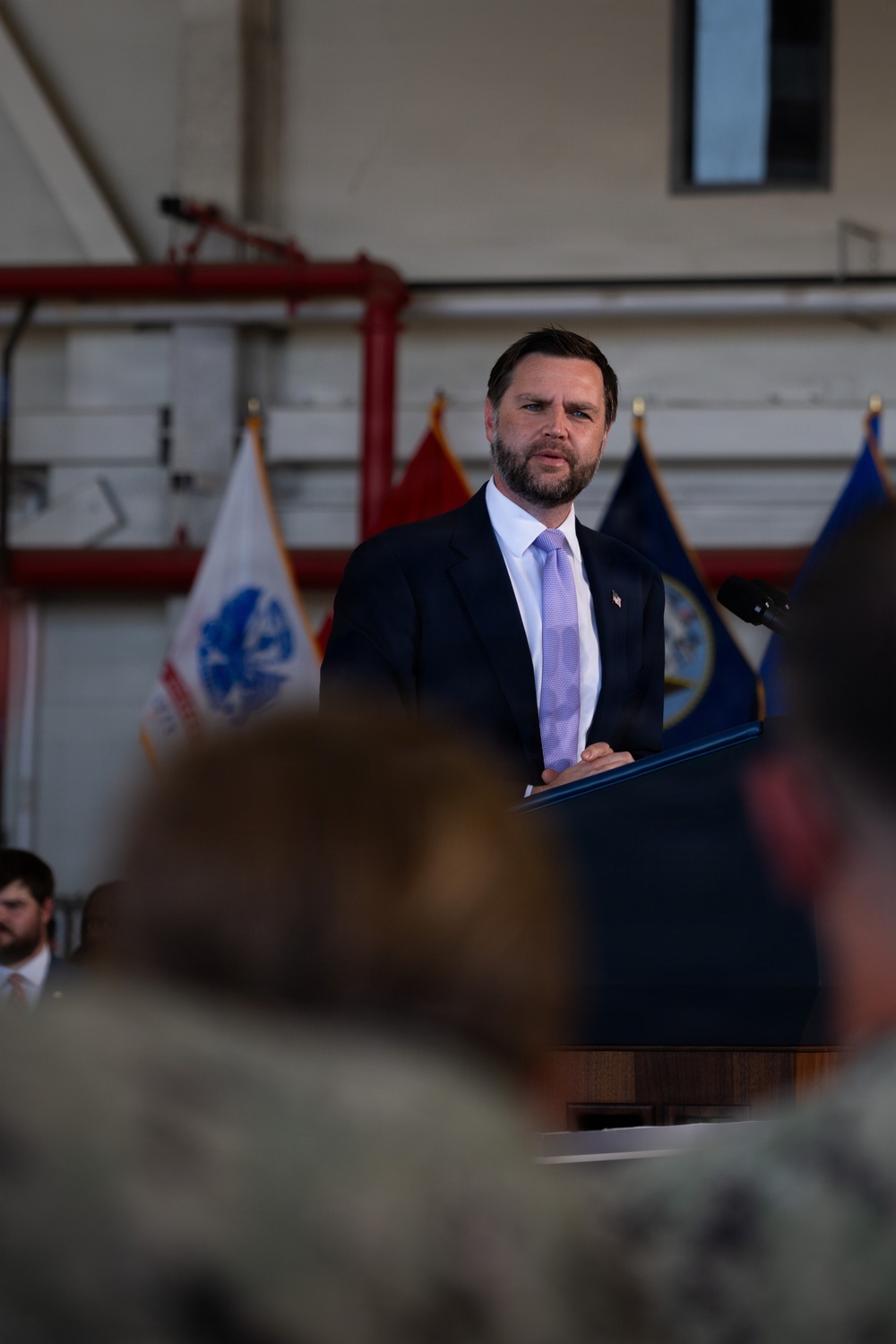 A man in a dark suit with a light purple tie stands at a wooden podium delivering a speech. The setting appears to be a military hangar or facility, with multiple flags visible in the background including what appears to be a U.S. Navy flag and other official flags. An audience, some in military camouflage uniforms, is visible in the foreground.
