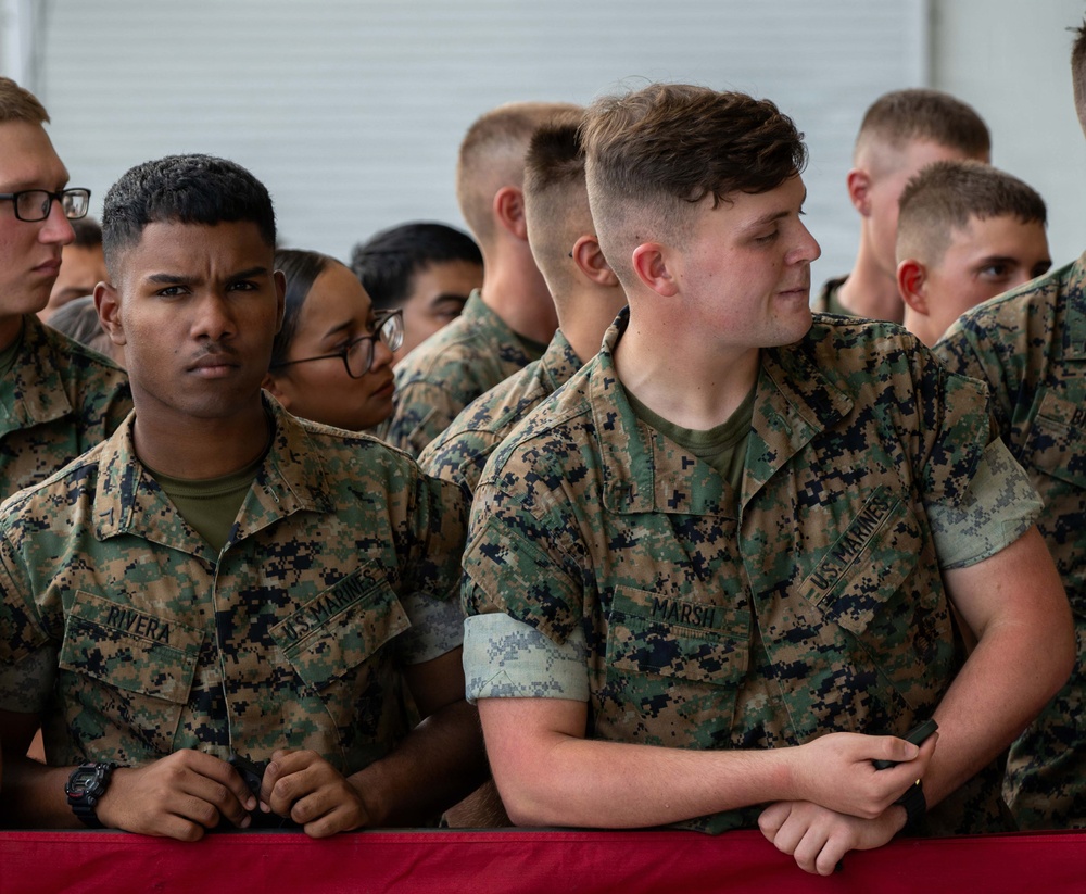 A group of U.S. Marines in digital camouflage uniforms (MARPAT) are gathered at what appears to be a formal military event or ceremony. Two Marines in the foreground are identifiable by their name tapes: 'Rivera' on the left and 'Marsh' on the right. They are standing behind a table draped with a red cloth, attentively observing proceedings.