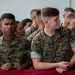 A group of U.S. Marines in digital camouflage uniforms (MARPAT) are gathered at what appears to be a formal military event or ceremony. Two Marines in the foreground are identifiable by their name tapes: 'Rivera' on the left and 'Marsh' on the right. They are standing behind a table draped with a red cloth, attentively observing proceedings.