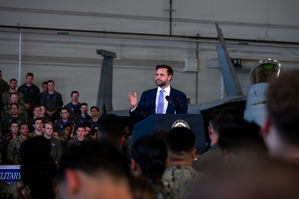 A man in a navy blue suit with a light blue tie is speaking at a podium inside a military hangar. The podium bears a partial seal reading 'ENT OF THE UNIT' (consistent with a Vice President or President of the United States seal). A large audience of military personnel in uniform fills the background, with a military aircraft visible behind the speaker.