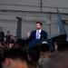A man in a navy blue suit with a light blue tie is speaking at a podium inside a military hangar. The podium bears a partial seal reading 'ENT OF THE UNIT' (consistent with a Vice President or President of the United States seal). A large audience of military personnel in uniform fills the background, with a military aircraft visible behind the speaker.