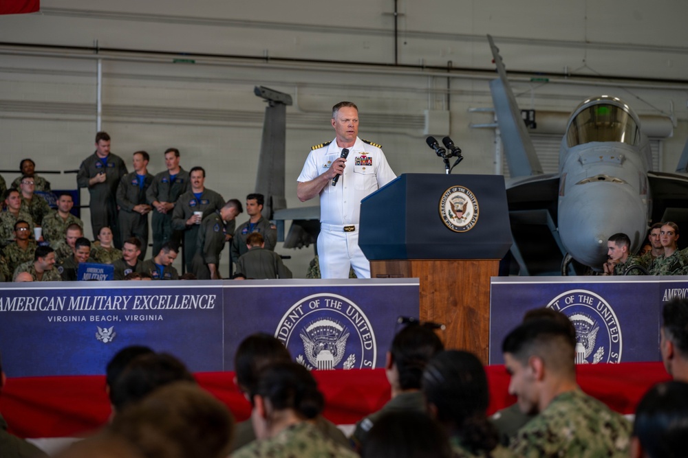 A U.S. Navy officer in white dress uniform speaks at a podium bearing the Vice President of the United States seal at an event titled 'American Military Excellence' in Virginia Beach, Virginia. The event is held inside a military hangar with military aircraft visible in the background. A large audience of military personnel in various uniforms is seated and standing in attendance.