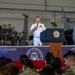 A U.S. Navy officer in white dress uniform speaks at a podium bearing the Vice President of the United States seal at an event titled 'American Military Excellence' in Virginia Beach, Virginia. The event is held inside a military hangar with military aircraft visible in the background. A large audience of military personnel in various uniforms is seated and standing in attendance.