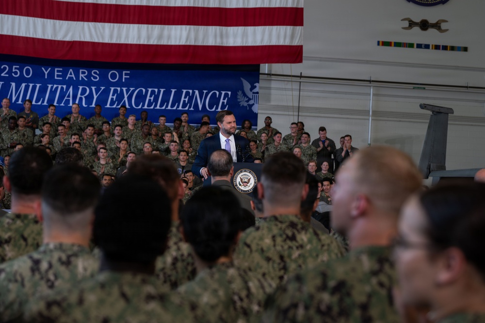 A civilian official in a dark suit is speaking at a podium bearing the Presidential Seal inside a military hangar, surrounded by a large audience of uniformed service members. A large American flag is draped in the background alongside a blue banner reading '250 Years of Military Excellence.' The setting suggests an official government address or ceremony at a U.S. military installation.