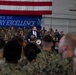 A civilian official in a dark suit is speaking at a podium bearing the Presidential Seal inside a military hangar, surrounded by a large audience of uniformed service members. A large American flag is draped in the background alongside a blue banner reading '250 Years of Military Excellence.' The setting suggests an official government address or ceremony at a U.S. military installation.