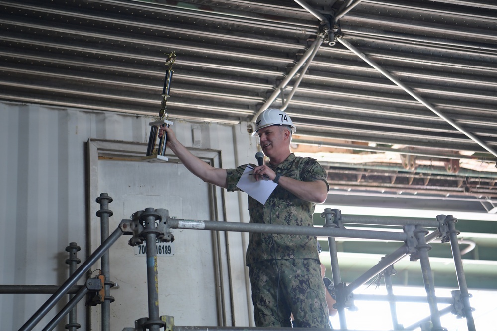 A military personnel member wearing a camouflage uniform and white hard hat marked '74' is holding up a trophy while standing on an elevated platform or scaffolding. The individual is also holding a microphone and papers, suggesting they are making an announcement or presentation. The setting appears to be an industrial or shipyard environment.