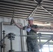 A military personnel member wearing a camouflage uniform and white hard hat marked '74' is holding up a trophy while standing on an elevated platform or scaffolding. The individual is also holding a microphone and papers, suggesting they are making an announcement or presentation. The setting appears to be an industrial or shipyard environment.