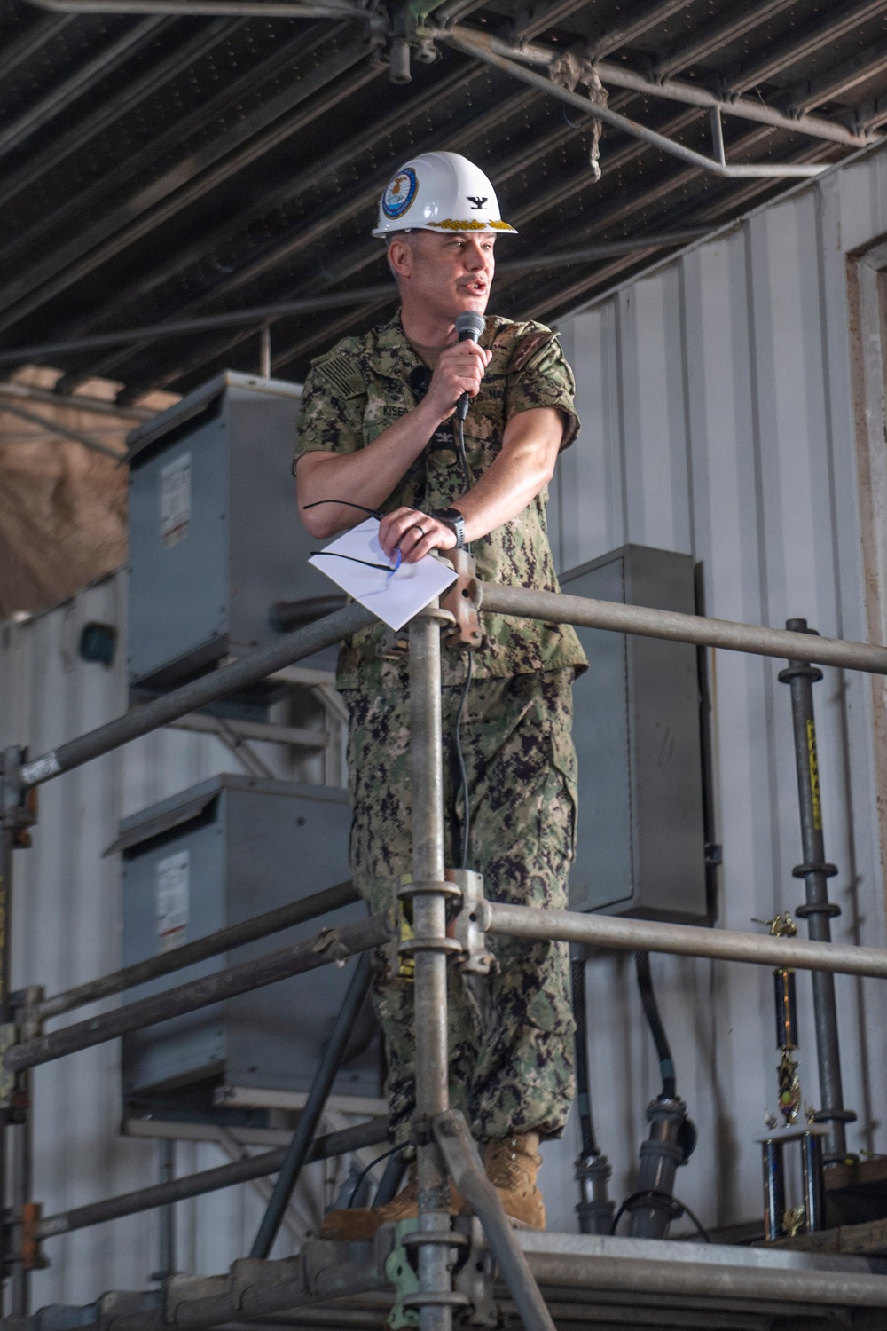 A U.S. Navy officer in camouflage utilities (NWU Type III) stands on scaffolding and speaks into a microphone while holding papers. He is wearing a white hard hat bearing a U.S. Navy seal and gold oak leaf cluster insignia indicating the rank of Captain (O-6). The setting appears to be an industrial or shipyard environment.