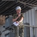 A U.S. Navy officer in camouflage utilities (NWU Type III) stands on scaffolding and speaks into a microphone while holding papers. He is wearing a white hard hat bearing a U.S. Navy seal and gold oak leaf cluster insignia indicating the rank of Captain (O-6). The setting appears to be an industrial or shipyard environment.
