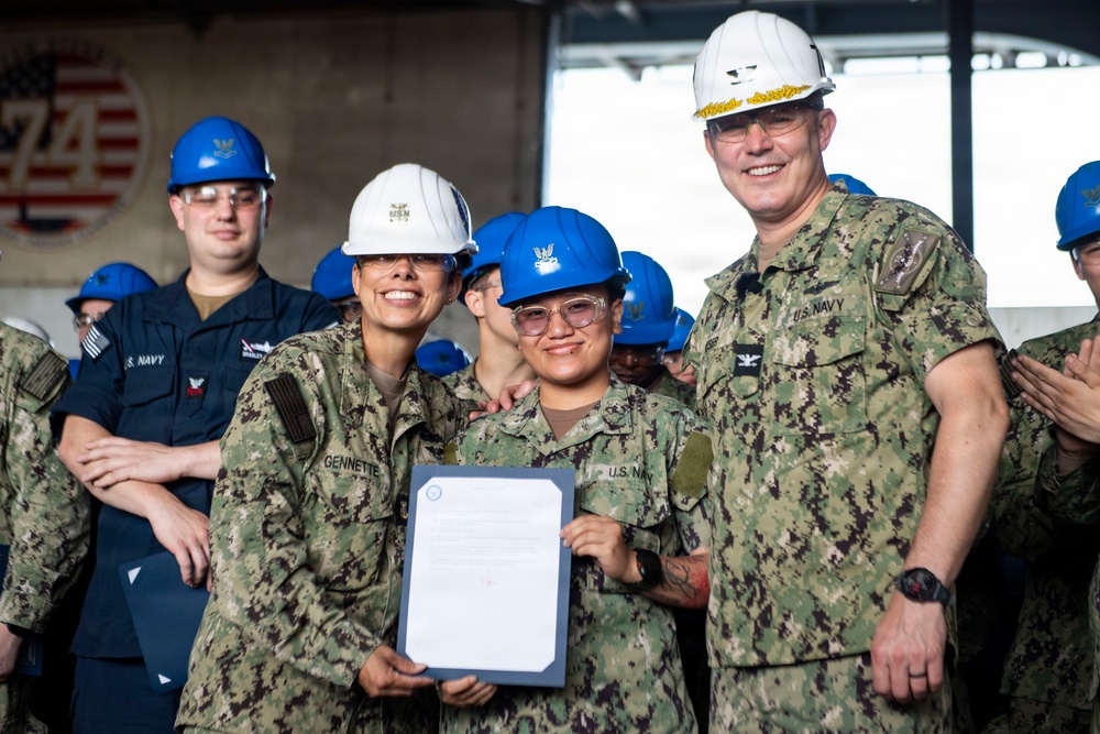 A U.S. Navy ceremony taking place in what appears to be a ship's hangar bay, where a sailor is being recognized with an official document. Three individuals are prominently featured in the foreground: two wearing white hard hats and one wearing a blue hard hat, all in Navy Working Uniform (NWU) camouflage. The background shows additional Navy personnel wearing blue hard hats and the number '74' with an American flag emblem visible on the wall.