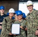 A U.S. Navy ceremony taking place in what appears to be a ship's hangar bay, where a sailor is being recognized with an official document. Three individuals are prominently featured in the foreground: two wearing white hard hats and one wearing a blue hard hat, all in Navy Working Uniform (NWU) camouflage. The background shows additional Navy personnel wearing blue hard hats and the number '74' with an American flag emblem visible on the wall.