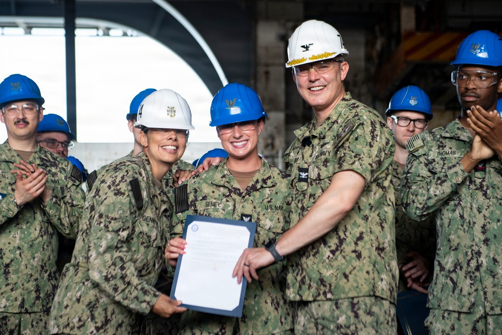 A group of U.S. Navy personnel in camouflage uniforms and hard hats are gathered for what appears to be a formal recognition or award ceremony. A senior officer wearing a white hard hat with a Captain's eagle insignia is presenting a document to a sailor identified by the name tape 'SENEY', while another sailor identified as 'GENI_' stands alongside. Surrounding personnel are applauding the occasion.