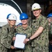 A group of U.S. Navy personnel in camouflage uniforms and hard hats are gathered for what appears to be a formal recognition or award ceremony. A senior officer wearing a white hard hat with a Captain's eagle insignia is presenting a document to a sailor identified by the name tape 'SENEY', while another sailor identified as 'GENI_' stands alongside. Surrounding personnel are applauding the occasion.