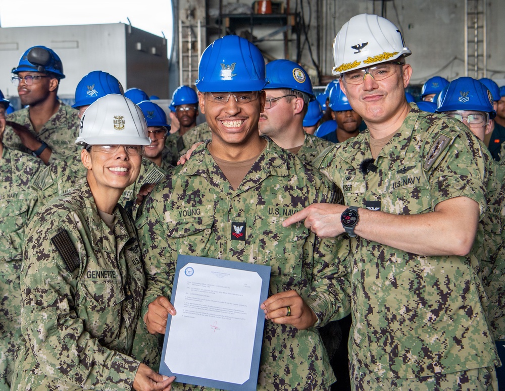 Three U.S. Navy personnel pose for a photo aboard what appears to be a naval vessel, with the central figure holding an official document. The individual in the center, identified by name tape as 'YOUNG,' is flanked by 'GENNETTE' on the left wearing a white hard hat and a senior officer on the right wearing a white hard hat with gold insignia. A large group of sailors wearing blue hard hats is visible in the background, suggesting a formal recognition or award ceremony.