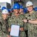 Three U.S. Navy personnel pose for a photo aboard what appears to be a naval vessel, with the central figure holding an official document. The individual in the center, identified by name tape as 'YOUNG,' is flanked by 'GENNETTE' on the left wearing a white hard hat and a senior officer on the right wearing a white hard hat with gold insignia. A large group of sailors wearing blue hard hats is visible in the background, suggesting a formal recognition or award ceremony.