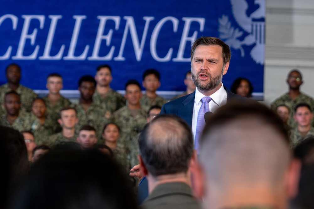 A man in a dark suit with a light purple tie is speaking at what appears to be a military event or address. He stands in front of a large blue banner partially reading 'CELLENCE' (likely 'EXCELLENCE') and a crowd of uniformed military personnel seated in rows behind him. The setting suggests an official ceremony or briefing at a military installation.