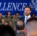 A man in a dark suit with a light purple tie is speaking at what appears to be a military event or address. He stands in front of a large blue banner partially reading 'CELLENCE' (likely 'EXCELLENCE') and a crowd of uniformed military personnel seated in rows behind him. The setting suggests an official ceremony or briefing at a military installation.