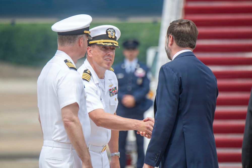 A formal military greeting ceremony is taking place on an airport tarmac near the stairs of an aircraft. Two U.S. Navy officers in white dress uniforms are shaking hands with a civilian man in a navy blue suit. The setting and protocol suggest an official arrival or departure ceremony.