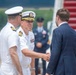 A formal military greeting ceremony is taking place on an airport tarmac near the stairs of an aircraft. Two U.S. Navy officers in white dress uniforms are shaking hands with a civilian man in a navy blue suit. The setting and protocol suggest an official arrival or departure ceremony.