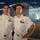 Two naval officers in white dress uniforms are posing together for a photograph, likely aboard a naval vessel or at a military event. Both men display rank insignia, medals, and qualification badges on their uniforms. A helicopter is visible in the background along with a projected aerial coastal image.