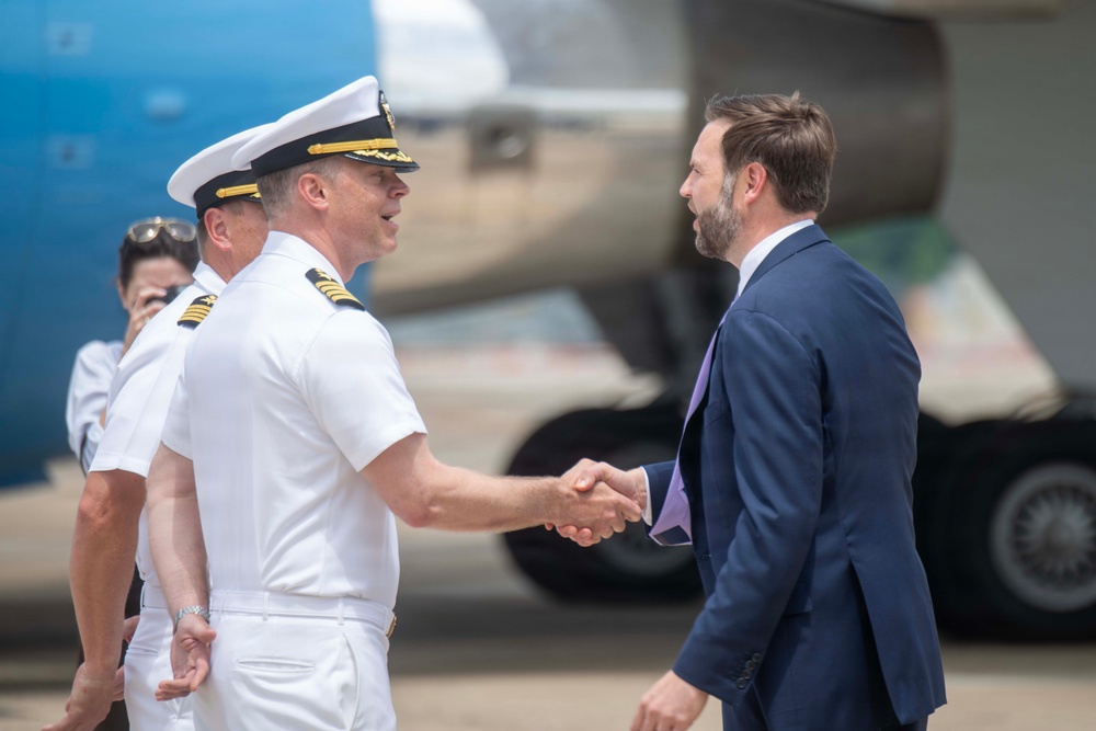 A formal greeting ceremony is taking place on an airport tarmac, where a naval officer in white dress uniform is shaking hands with a civilian man in a navy blue suit. A large blue and white aircraft is visible in the background, suggesting an official arrival or departure event. Additional personnel in white uniforms are visible in the background.