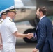 A formal greeting ceremony is taking place on an airport tarmac, where a naval officer in white dress uniform is shaking hands with a civilian man in a navy blue suit. A large blue and white aircraft is visible in the background, suggesting an official arrival or departure event. Additional personnel in white uniforms are visible in the background.