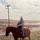 A vintage photograph showing an adult and a young child riding together on a chestnut/sorrel horse in an open, arid rangeland setting. The adult, wearing a blue jacket and baseball cap, holds the child securely in front of them on the saddle. A wooden utility pole with power lines is visible in the background against a flat, semi-arid landscape.