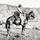 A black-and-white photograph depicting a cowboy on horseback in an open rangeland setting with rolling hills in the background. The rider is equipped with traditional Western gear including a wide-brimmed hat, chaps, and a coiled lasso. The image appears to date from the early-to-mid 20th century based on photographic quality and style.