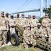 A group of approximately 13 U.S. Army soldiers in MultiCam (OCP) camouflage uniforms pose for a group photo outdoors. They are positioned around what appears to be a historic artillery piece, with a large suspension bridge visible in the background. The setting appears to be a military installation or park near a major waterway.