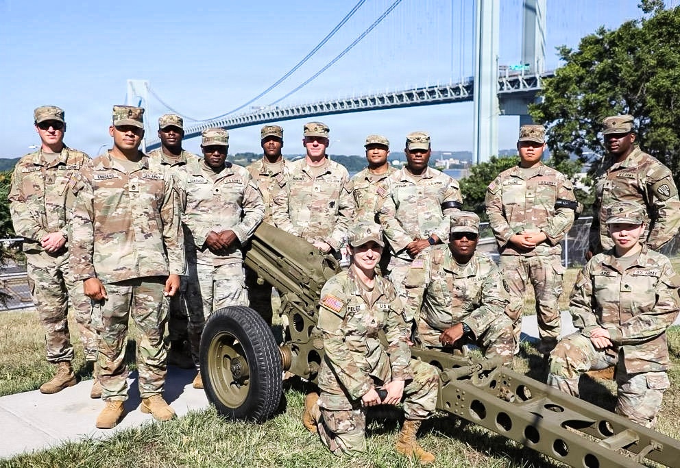 A group of approximately 13 U.S. Army soldiers in MultiCam (OCP) camouflage uniforms pose for a group photo outdoors. They are positioned around what appears to be a historic artillery piece, with a large suspension bridge visible in the background. The setting appears to be a military installation or park near a major waterway.