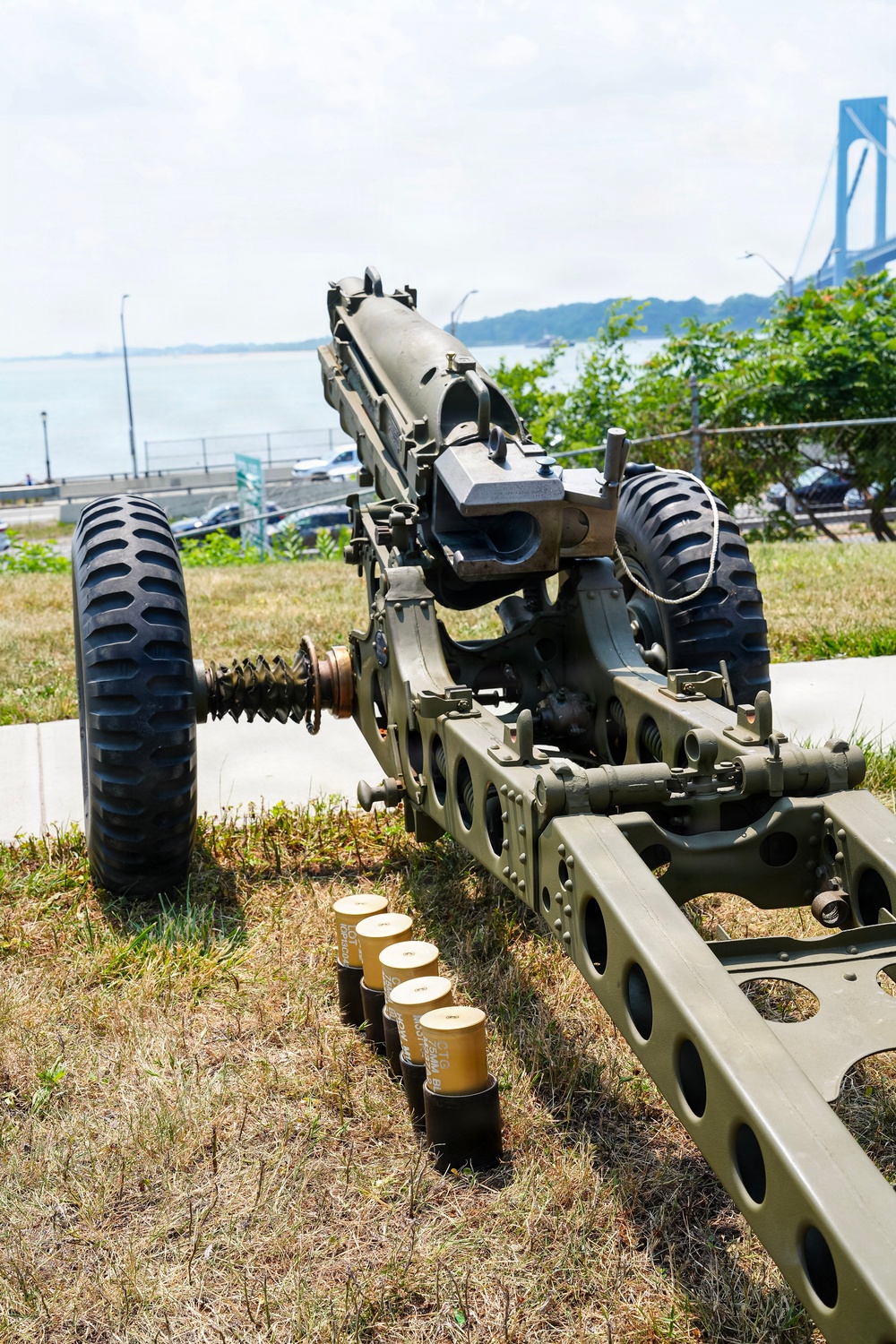 A military artillery piece, likely a howitzer or field gun, is displayed outdoors on a grassy area near a waterfront. Several artillery rounds/shells are arranged on the ground in front of the weapon. A large suspension bridge, consistent with the Verrazzano-Narrows Bridge in New York, is visible in the background.