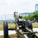 A military artillery piece, likely a howitzer or field gun, is displayed outdoors on a grassy area near a waterfront. Several artillery rounds/shells are arranged on the ground in front of the weapon. A large suspension bridge, consistent with the Verrazzano-Narrows Bridge in New York, is visible in the background.