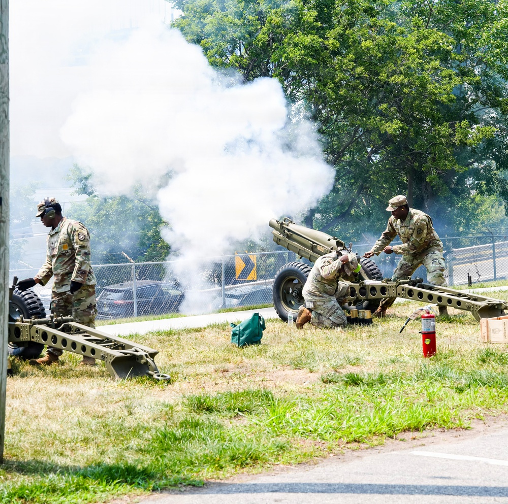 Three soldiers in U.S. Army OCP (Operational Camouflage Pattern) uniforms are operating a light artillery piece, likely an M119 howitzer or ceremonial cannon, which has just been fired as evidenced by the large white smoke cloud billowing from the muzzle. The scene appears to be a ceremonial salute or demonstration rather than combat, given the urban/base setting, proximity to a road, and presence of a fire extinguisher nearby as a safety precaution. The soldiers are positioned around the cannon in post-firing stances.