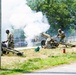 Three soldiers in U.S. Army OCP (Operational Camouflage Pattern) uniforms are operating a light artillery piece, likely an M119 howitzer or ceremonial cannon, which has just been fired as evidenced by the large white smoke cloud billowing from the muzzle. The scene appears to be a ceremonial salute or demonstration rather than combat, given the urban/base setting, proximity to a road, and presence of a fire extinguisher nearby as a safety precaution. The soldiers are positioned around the cannon in post-firing stances.