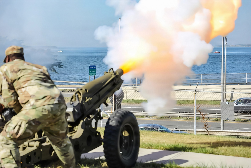 A U.S. Army soldier in MultiCam/OCP camouflage uniform is firing a ceremonial artillery cannon, producing a large muzzle blast with orange flame and white smoke. The scene appears to be a formal military ceremony conducted near a waterfront road, likely at a military installation. Civilian vehicles and a body of water with boats are visible in the background.