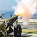 A U.S. Army soldier in MultiCam/OCP camouflage uniform is firing a ceremonial artillery cannon, producing a large muzzle blast with orange flame and white smoke. The scene appears to be a formal military ceremony conducted near a waterfront road, likely at a military installation. Civilian vehicles and a body of water with boats are visible in the background.