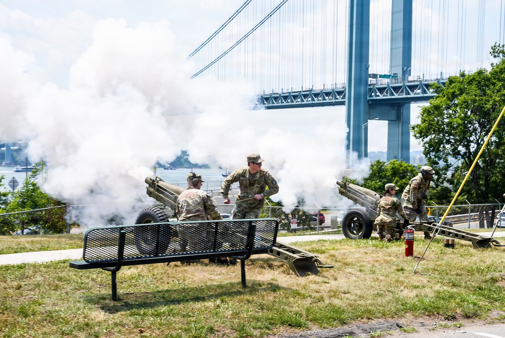 Military personnel in camouflage uniforms are operating artillery cannons in an outdoor park setting, producing large clouds of white smoke consistent with a ceremonial salute firing. The scene takes place near a large suspension bridge, identifiable as the Verrazzano-Narrows Bridge in New York. A park bench and fire extinguisher are visible in the foreground, suggesting a public ceremonial event.