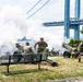 Military personnel in camouflage uniforms are operating artillery cannons in an outdoor park setting, producing large clouds of white smoke consistent with a ceremonial salute firing. The scene takes place near a large suspension bridge, identifiable as the Verrazzano-Narrows Bridge in New York. A park bench and fire extinguisher are visible in the foreground, suggesting a public ceremonial event.