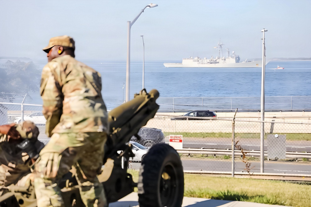 A soldier in camouflage uniform and wearing ear protection stands near an artillery piece, likely a howitzer, positioned on a grassy area near a waterfront. In the background, a large naval vessel is visible on the water, along with a road, chain-link fence, and street lights. The scene suggests a military ceremony or training exercise at a coastal military installation.