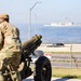 A soldier in camouflage uniform and wearing ear protection stands near an artillery piece, likely a howitzer, positioned on a grassy area near a waterfront. In the background, a large naval vessel is visible on the water, along with a road, chain-link fence, and street lights. The scene suggests a military ceremony or training exercise at a coastal military installation.