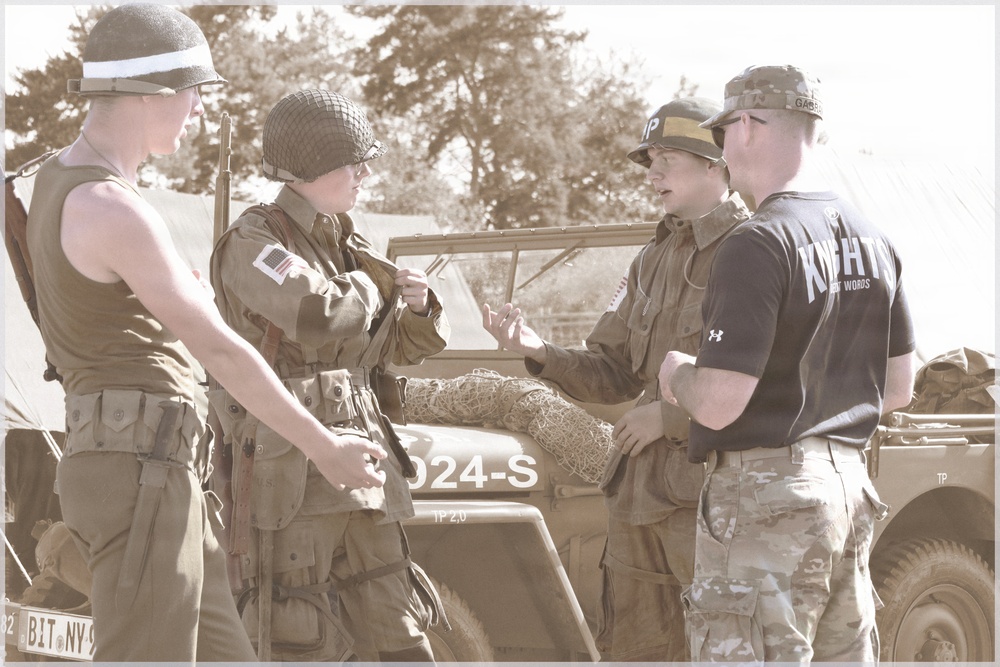 The image shows four individuals gathered around a military jeep, with two wearing WWII-era American uniforms and helmets, one in modern multicam military pants with a black t-shirt, and one in a 'MP' cap with period uniform. The vehicle bears the marking '024-S' and appears to be a WWII-era Willys jeep, suggesting this is a military reenactment event. The group appears to be engaged in conversation.