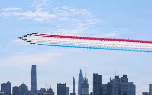 A formation of military jets performs an aerobatic flyover above the Manhattan skyline, releasing red, white, and blue smoke trails in a patriotic display. The scene is viewed from across the Hudson River, with a naval warship visible in the foreground water. The iconic Empire State Building and other recognizable Midtown Manhattan skyscrapers are clearly visible in the background.